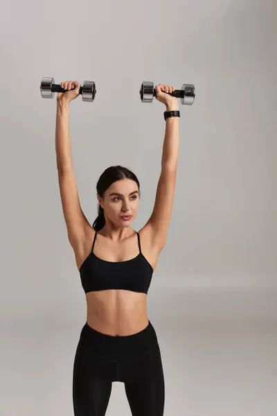 Femme forte et sportive en noir active porter haltères de levage au-dessus de la tête sur fond gris — Photo de stock