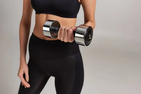 Cropped shot of slim sportswoman in active wear lifting dumbbell while working out on grey backdrop — Stock Photo