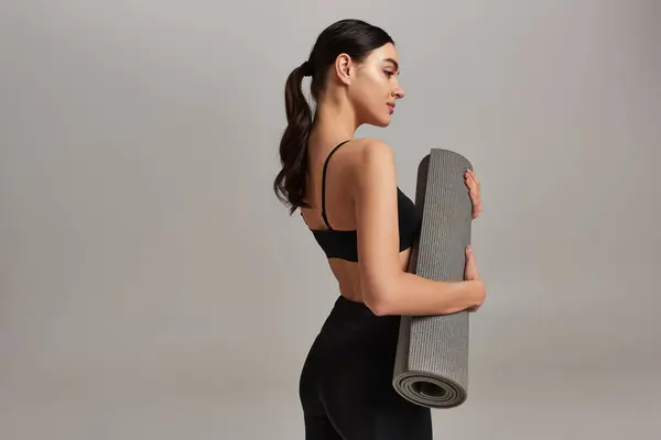 Side view, sportswoman in crop top and leggings holding fitness mat and standing on grey background — Stock Photo