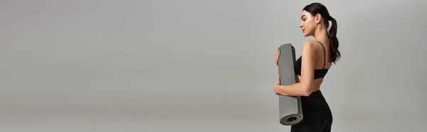 Вид збоку спортсменки у верхній частині врожаю і легінси, що тримають фітнес-матер і стоять на сірому, банер — стокове фото