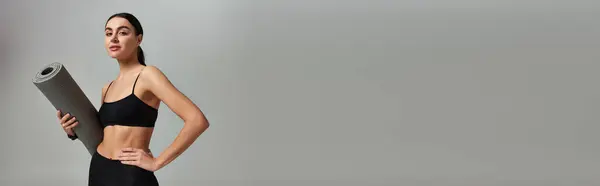 Esportista em desgaste ativo segurando tapete de fitness e de pé com a mão no quadril em cinza, banner — Fotografia de Stock