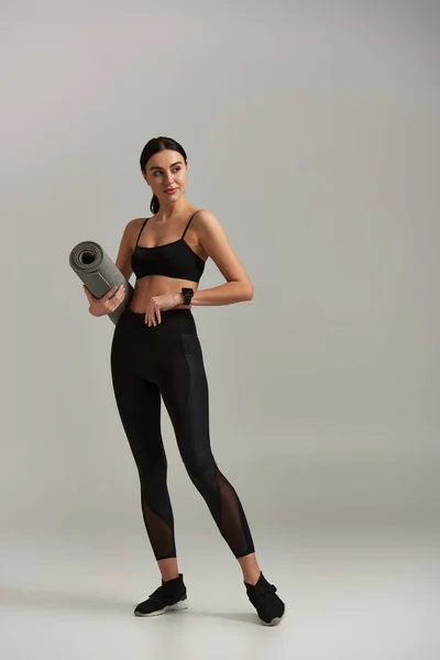 Volle Länge der fitten Sportlerin in aktiver Kleidung hält Fitnessmatte und steht auf grauem Hintergrund — Stockfoto