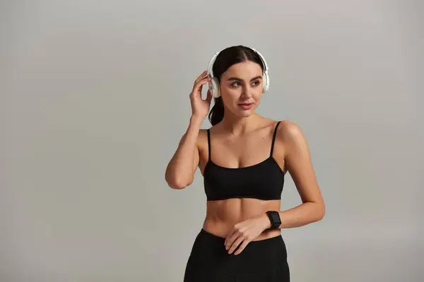 Pretty young sportswoman in crop top listening music in wireless headphones on grey background — Stock Photo