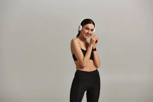 Довольная спортсменка в активной одежде слушает музыку в беспроводных наушниках на сером фоне — стоковое фото