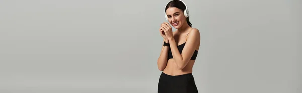 Pleased sportswoman in active wear listening music in wireless headphones on grey background, banner — Stock Photo