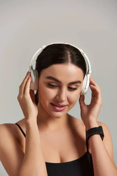 Zufriedene Sportlerin in aktiver Kleidung hört Musik und berührt drahtlose Kopfhörer vor grauem Hintergrund — Stockfoto