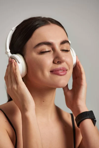Щаслива спортсменка в активному носінні слухає музику і торкається бездротових навушників на сірому фоні — стокове фото