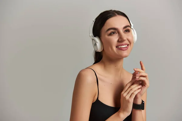 Mujer joven feliz en desgaste activo escuchando música en auriculares inalámbricos sobre fondo gris - foto de stock