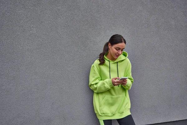 Молодая женщина в лаймовом цвете толстовка и леггинсы сообщения на смартфоне рядом с серой бетонной стеной — стоковое фото