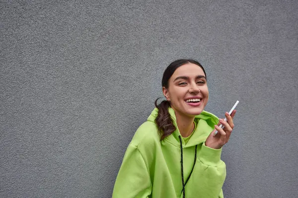 Cheerful woman in lime color hoodie holding smartphone and smiling near grey concrete wall — Stock Photo