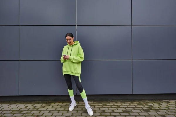 Femme heureuse en couleur citron vert sweat à capuche et leggings messagerie sur smartphone près du mur gris à l'extérieur — Photo de stock