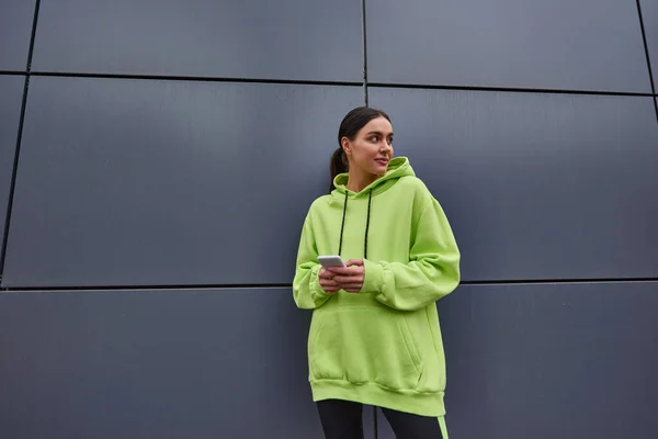 Joyful woman in lime color hoodie holding smartphone and looking away while standing near grey wall — Stock Photo