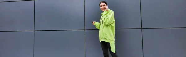 Щаслива жінка в лаймовому кольорі светр і легінси розмовляють на смартфоні біля сірої стіни на відкритому повітрі, банер — стокове фото