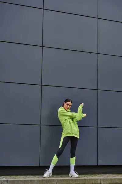 Bonita deportista con capucha y polainas calentándose cerca de la pared gris al aire libre, aire fresco y deporte - foto de stock