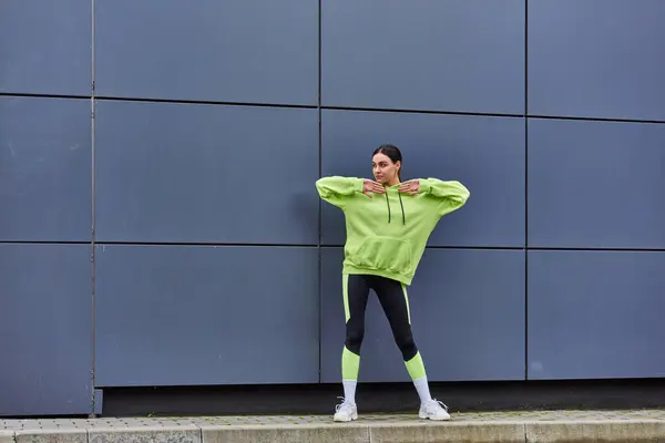 Jeune sportive en sweat à capuche et leggings échauffant près du mur gris à l'extérieur, de l'air frais et du sport — Photo de stock