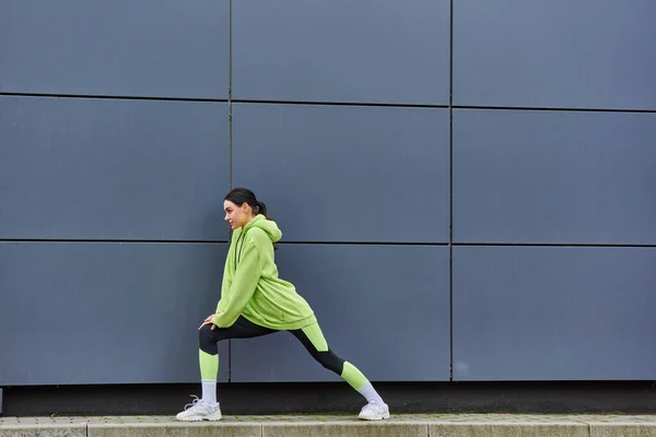 Jeune sportive en sweat à capuche et leggings étirant les jambes près du mur gris à l'extérieur, l'énergie et le sport — Photo de stock