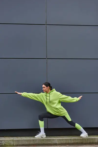 Jeune sportive en tenue de sport avec les mains tendues étirant les jambes près du mur gris à l'extérieur — Photo de stock