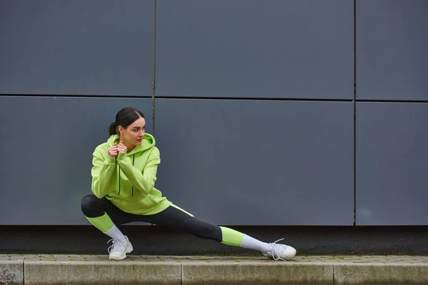 Jovem desportista de capuz e leggings fazendo lunges perto de parede cinza ao ar livre, energia e esporte — Fotografia de Stock
