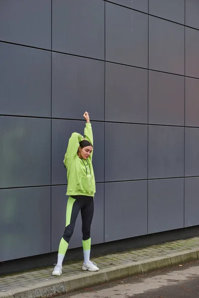 Joven deportista con capucha y polainas calentándose cerca de la pared gris al aire libre, motivación y deporte - foto de stock