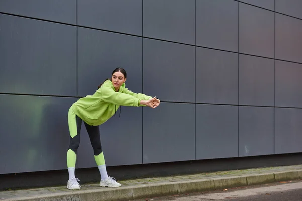 Атлетичная женщина в толстовке и леггинсах разогревается возле серой стены на открытом воздухе, мотивации и спорте — стоковое фото