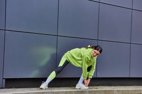 Mulher enérgica em capuz e leggings aquecimento perto de parede cinza ao ar livre, motivação e esporte — Fotografia de Stock