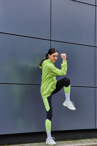 Atlético feminino corredor em capuz e leggings aquecimento perto de cinza parede ao ar livre, motivação — Fotografia de Stock