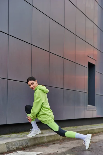 Bonita corredora en sudadera con capucha y polainas haciendo embestidas cerca de la pared gris al aire libre, motivación - foto de stock