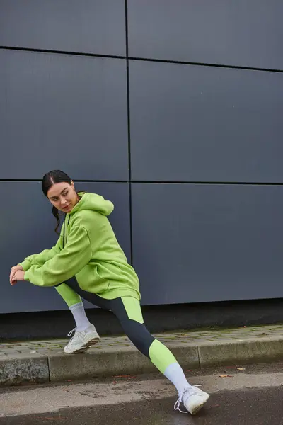 Sportive en sweat à capuche de couleur lime et leggings étirant les jambes près du mur gris à l'extérieur, motivation — Photo de stock
