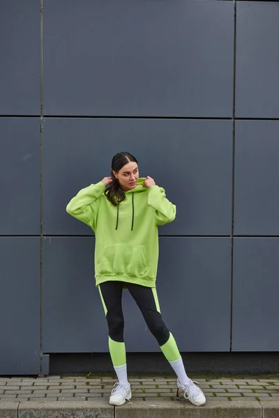 Attractive sportswoman in lime color hoodie and leggings standing near grey wall outdoors, sport — Stock Photo