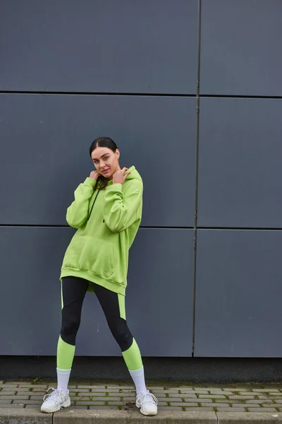Beautiful sportswoman in lime color hoodie and leggings standing near grey wall outdoors, sport — Stock Photo