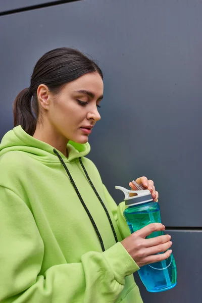 Young brunette sportswoman in lime color hoodie holding bottle of water near grey wall outdoors — Stock Photo