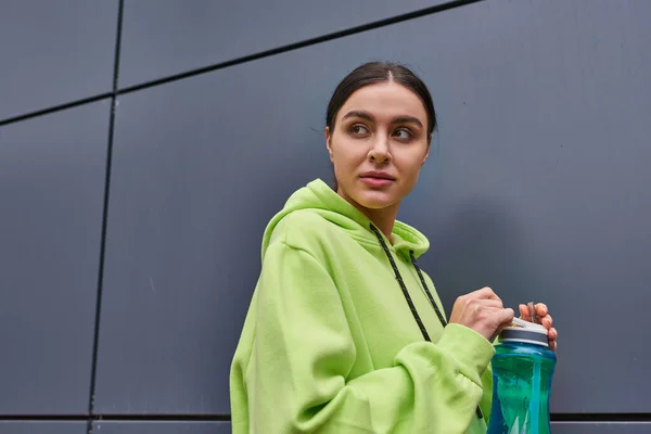 Junge attraktive Sportlerin in kalkfarbenem Kapuzenpulli mit Wasserflasche an der grauen Wand im Freien — Stockfoto