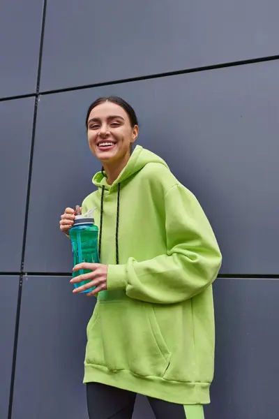 Deportista feliz y joven en color lima sudadera con capucha celebración botella de agua cerca de la pared gris al aire libre - foto de stock