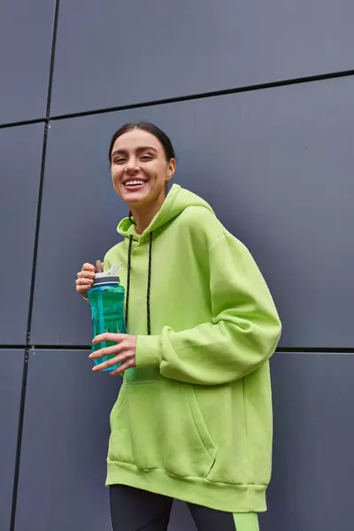 Happy and young sportswoman in lime color hoodie holding bottle of water near grey wall outdoors — Stock Photo