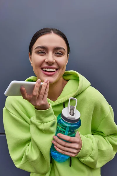 Cheerful sportswoman in hoodie holding bottle of water and recording audio message on smartphone — Stock Photo