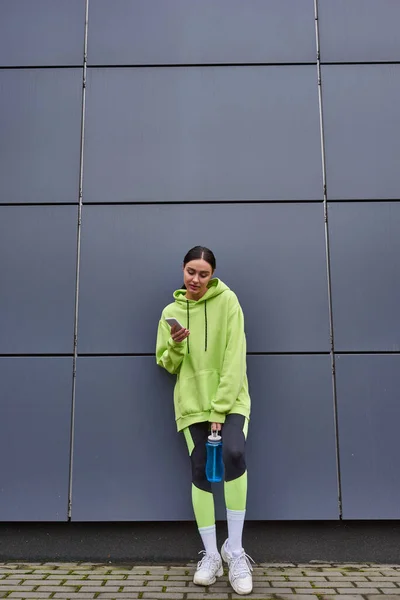 Pretty sportswoman in hoodie holding bottle of water and using smartphone near grey wall outdoors — Stock Photo