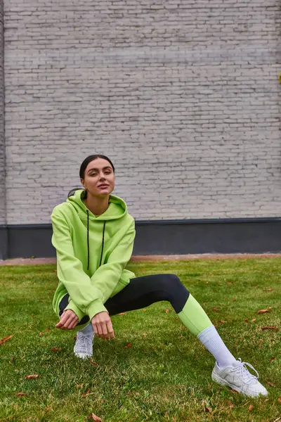 Stylish sportswoman in lime color hoodie and leggings sitting on green grass, healthy lifestyle — Stock Photo