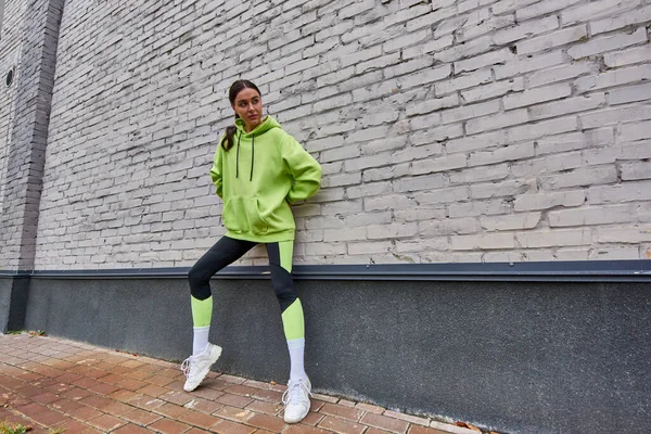 Stylish young sportswoman in lime color hoodie, leggings and sneakers standing near grey wall — Stock Photo