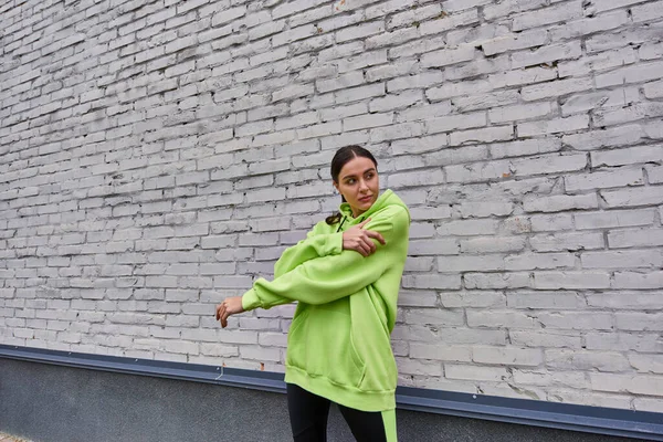 Attraktive junge Sportlerin in lindfarbenem Kapuzenpulli und Leggings, die sich in der Nähe der grauen Wand im Freien ausstrecken — Stockfoto