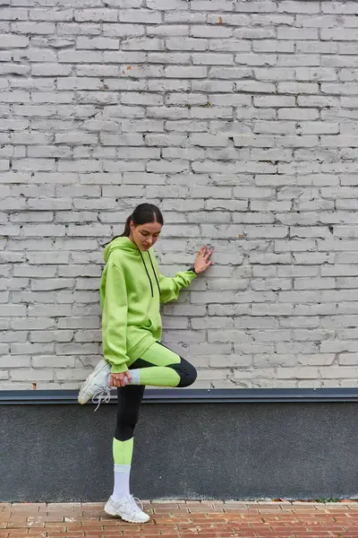 Pretty young sportswoman in lime color hoodie and leggings stretching leg near grey wall outdoors — Stock Photo