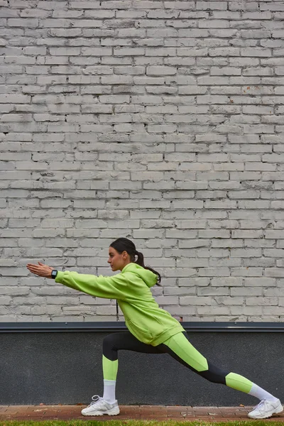 Vista lateral de la deportista en sudadera con capucha color lima y polainas haciendo saltos antes de correr al aire libre - foto de stock