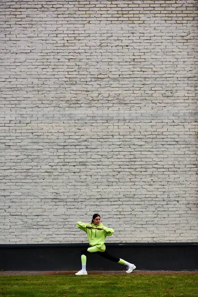 Deportista activa en sudadera con capucha de color lima y polainas haciendo embestidas mientras hace ejercicio al aire libre - foto de stock