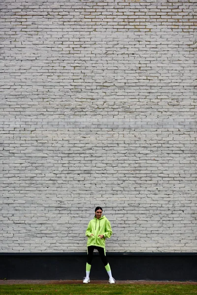 Pretty sportswoman in lime color hoodie and leggings standing with hands in pockets near brick wall — Stock Photo