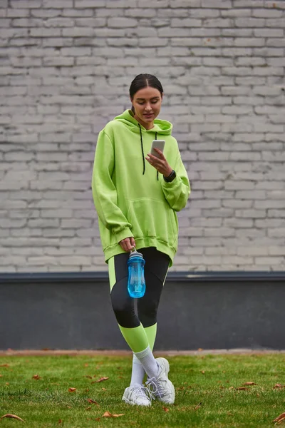 Brunette sportswoman in hoodie and leggings using smartphone and standing with bottle with water — Stock Photo