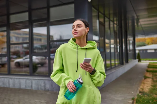 Sportswoman in lime color hoodie using smartphone and standing with bottle with water outdoors — Stock Photo