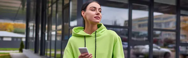 Спортсменка в лаймовом цвете толстовка с помощью смартфона и стоя рядом с современным зданием, баннер — стоковое фото