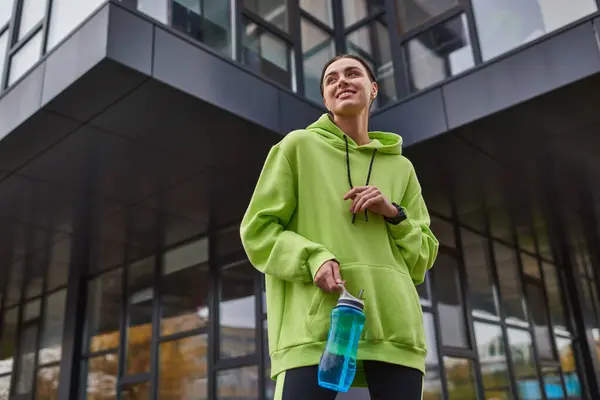 Glückliche Sportlerin in überdimensionalem Kapuzenpulli und Leggings, die mit Flasche mit Wasser im Freien steht — Stockfoto