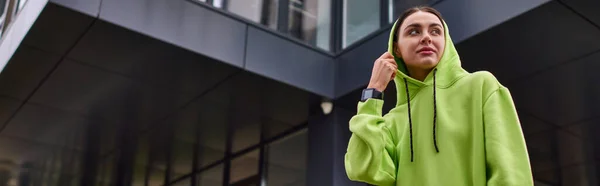 Banner einer jungen Sportlerin mit Kapuze auf dem Kopf, die in der Nähe eines modernen Gebäudes steht, lindfarbener Kapuzenpullover — Stockfoto