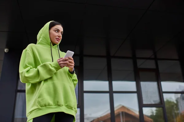 Junge brünette Frau mit Kapuze auf dem Kopf mit Smartphone in der Nähe moderner Gebäude, Street-Style-Look — Stockfoto