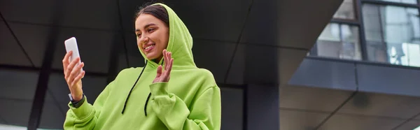 Mujer joven feliz en sudadera con capucha saludando la mano mientras tiene videollamada en el teléfono inteligente cerca del edificio, pancarta - foto de stock